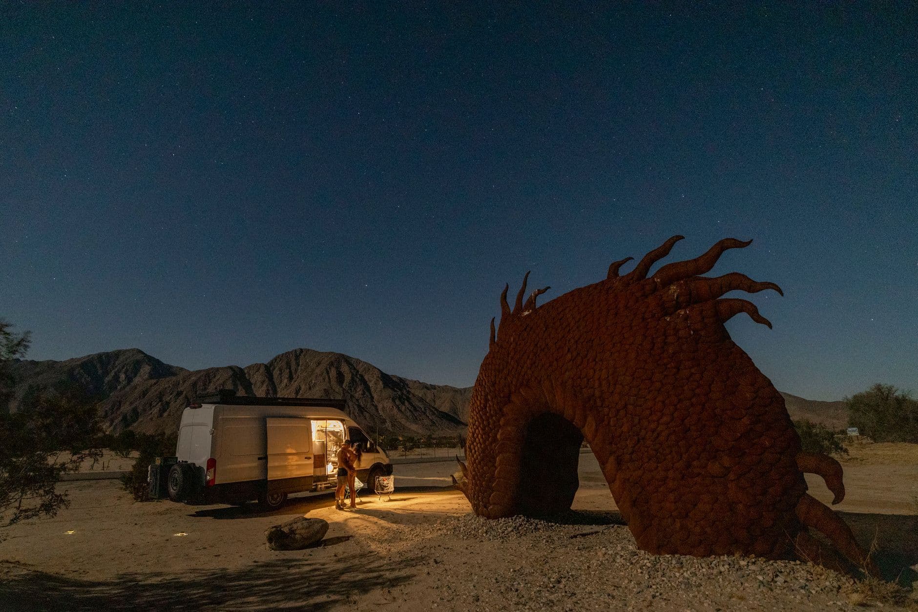 Camping with a van beside a dragon statue under a starry desert sky at night.
