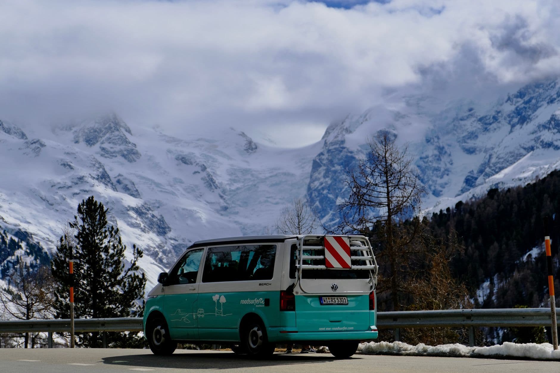 A scenic view of a teal van parked on a road with majestic snow-covered mountains in the background.