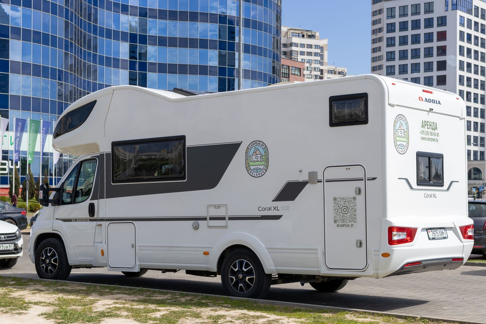 A white RV parked beside modern glass buildings in a sunny urban setting.