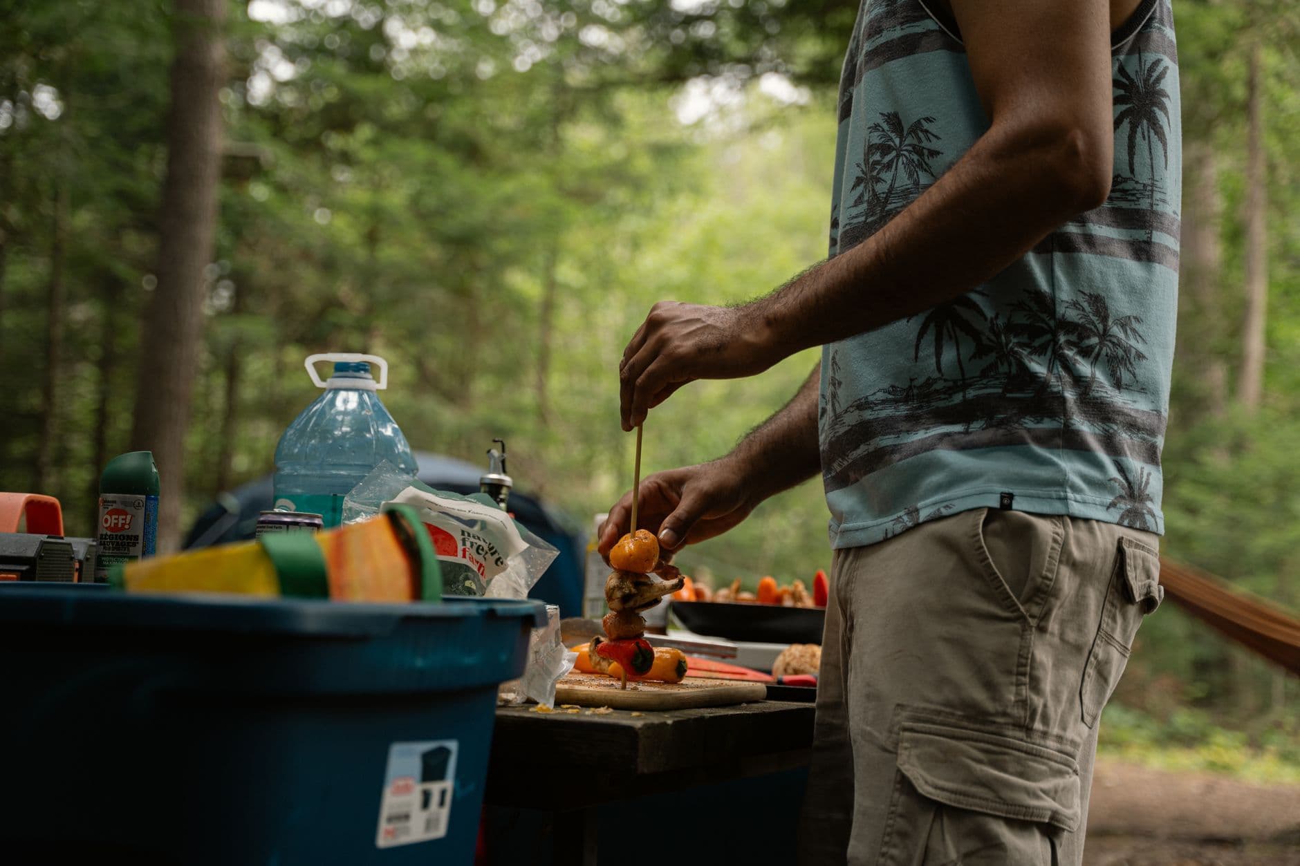 Adult man assembling skewers at a camp kitchen surrounded by forest during the day.