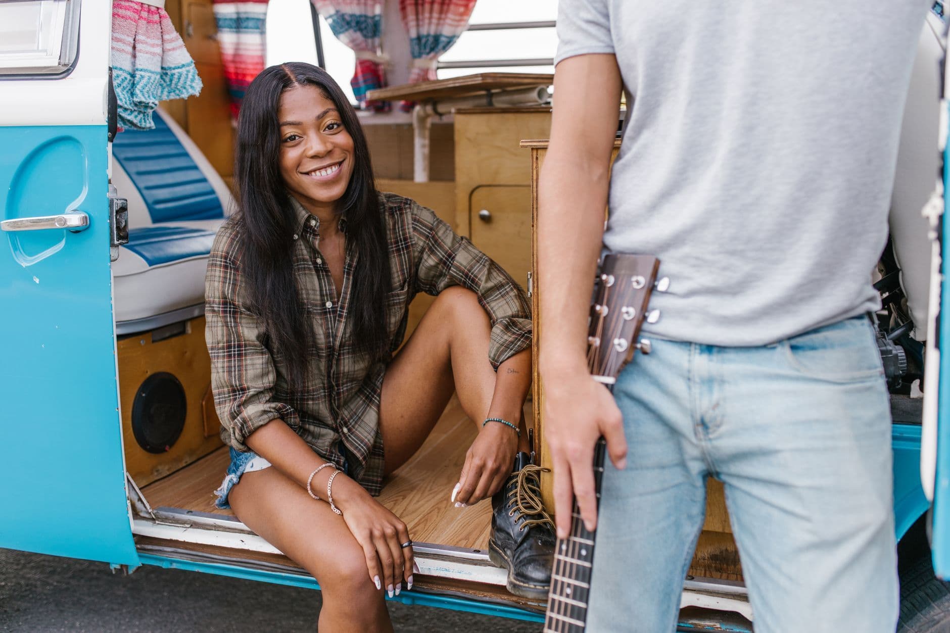 A cheerful woman sits in a campervan door while another person holds a guitar nearby.