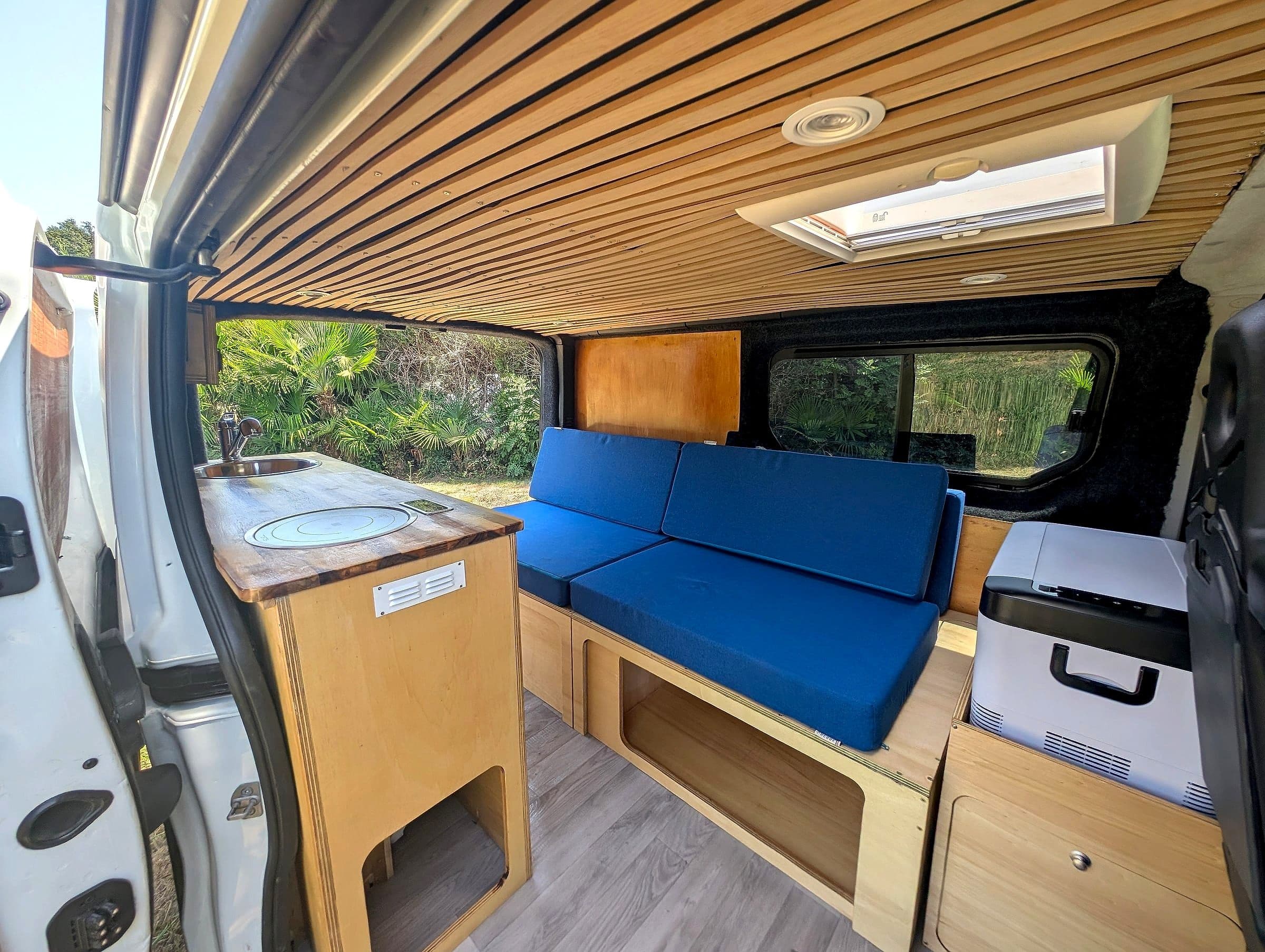 Intérieur moderne et lumineux d'un van aménagé Vanzon Explorer avec cuisine en bois, banquettes bleues et vue sur la nature.