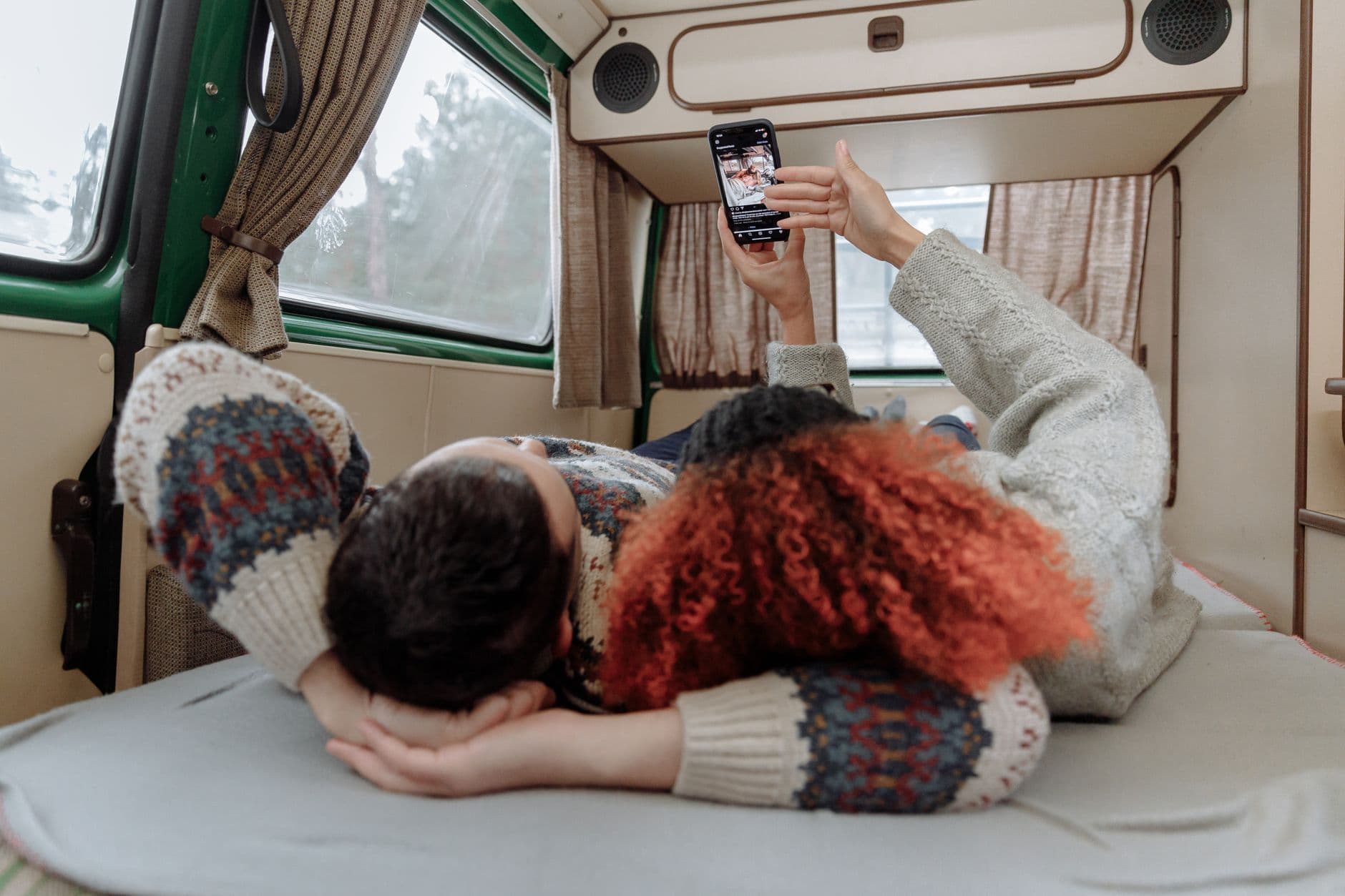 A couple lounging inside a cozy camper van, watching videos on a smartphone.